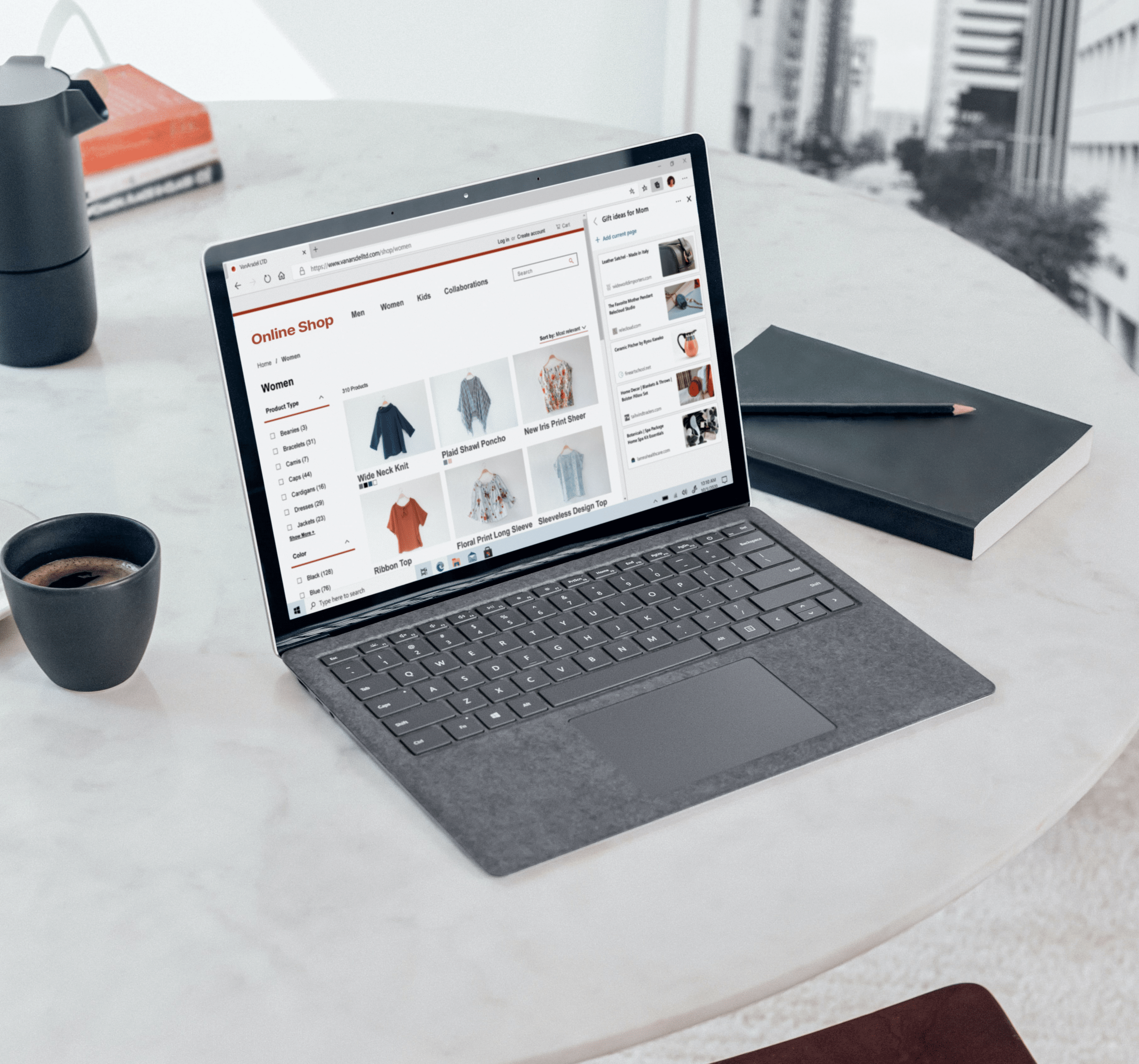A laptop displaying an online clothing shop sits on a marble table next to a coffee cup, a notebook, a pencil, and a modern kettle, suggesting a work or shopping setup.