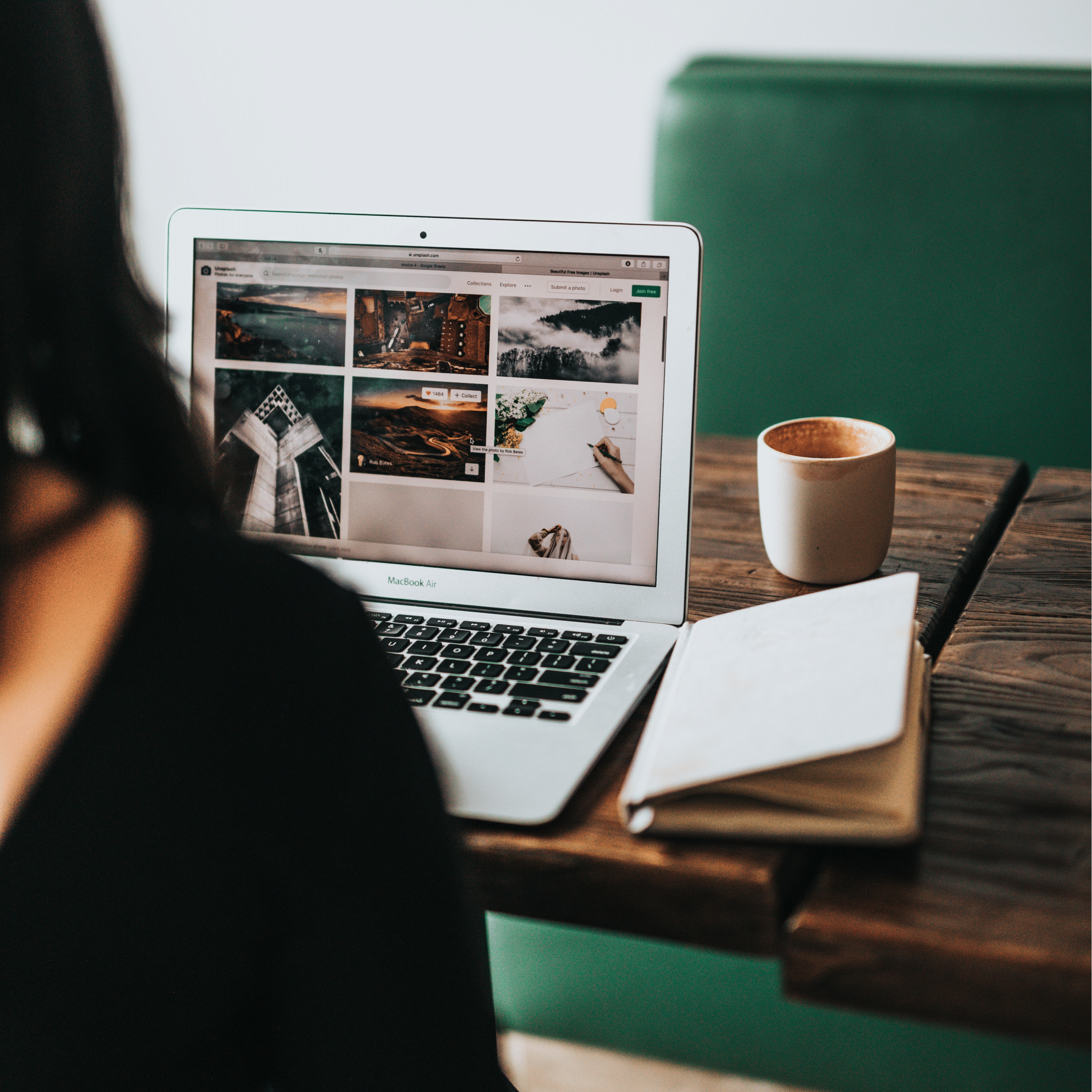 A person sitting at a wooden table, while working on a laptop computer, searching for images to include on the SmartHost SiteJet Website Builder. A cup of coffee and a notebook are also on the table