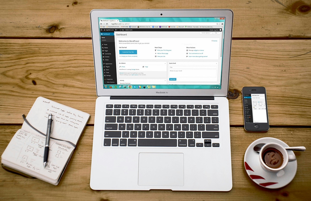 A laptop displaying a website dashboard is on a wooden table. Nearby are a notebook, pen, smartphone, and a cup of coffee.