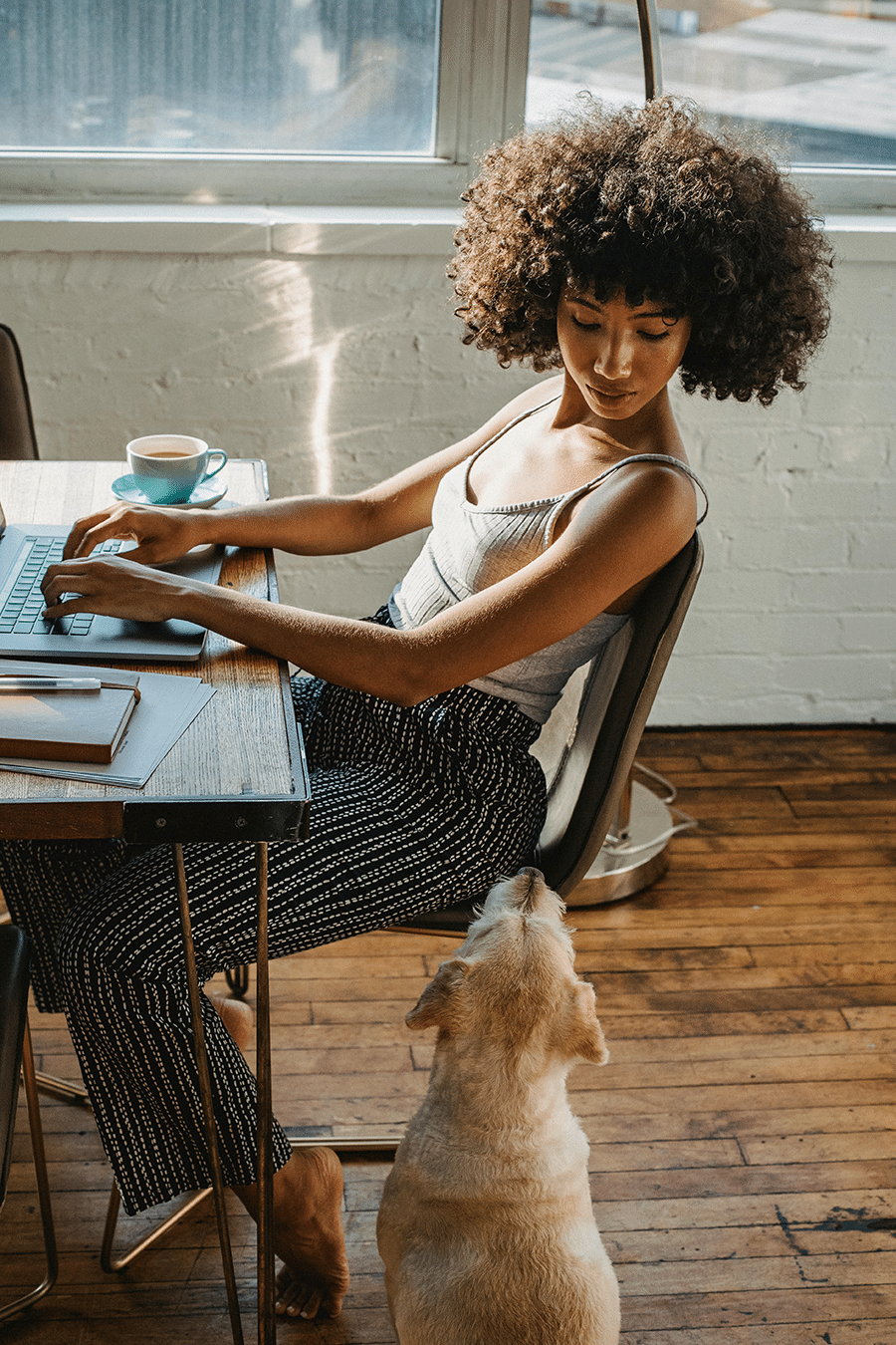 A woman types on a laptop at a desk while looking down at a dog sitting beside her on a wooden floor.