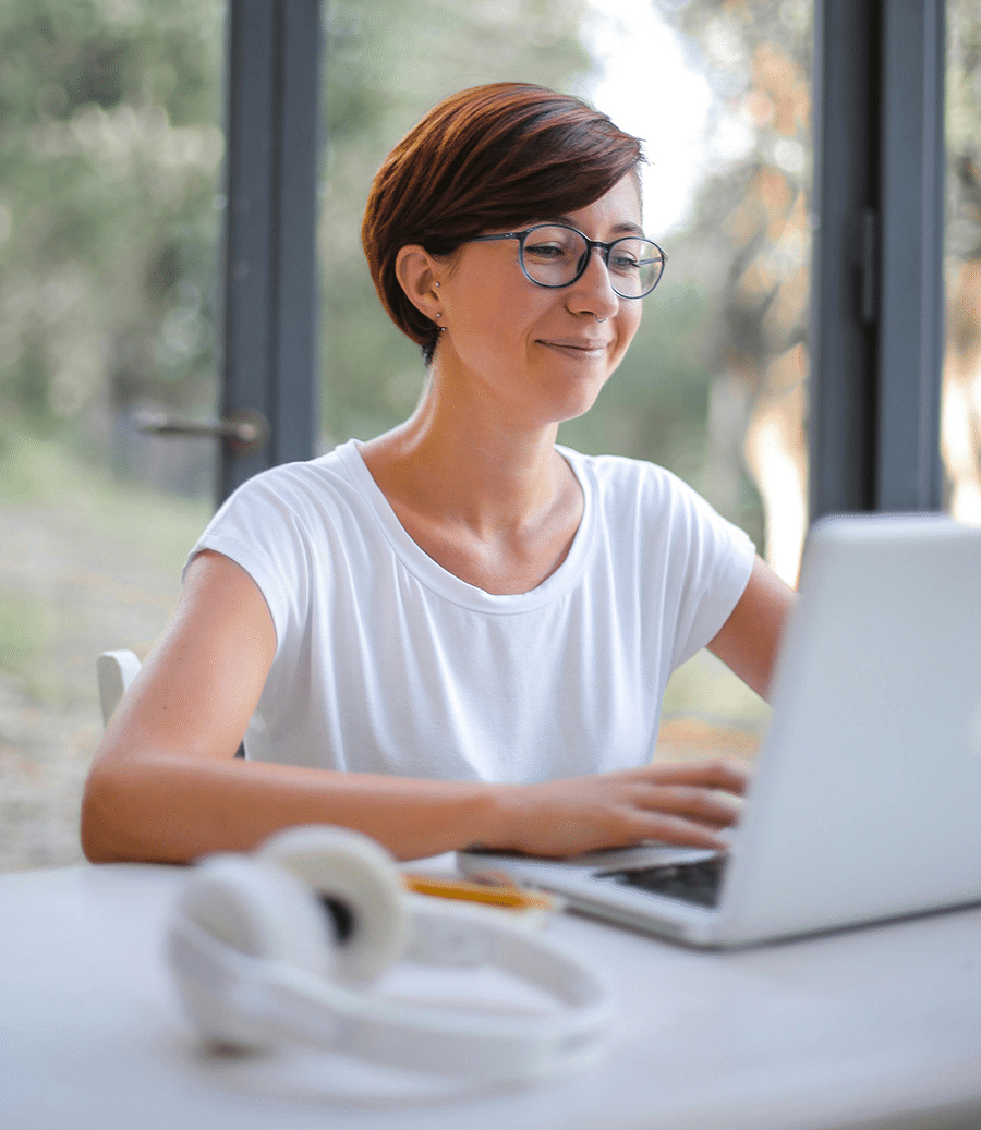 Woman with short brown hair and glasses smiles while working on a laptop at a desk, with headphones and a pencil nearby, in a bright room with large windows.