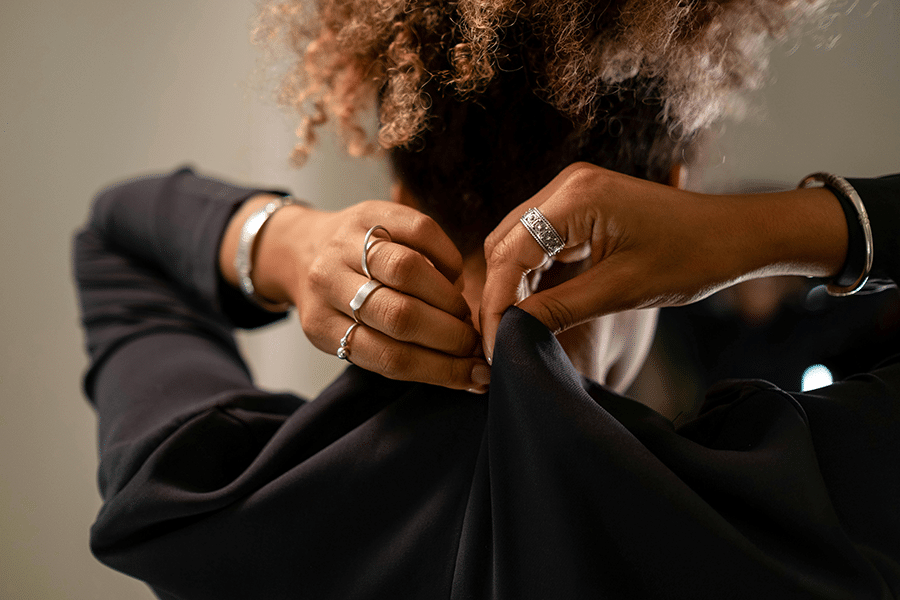 A person with curly hair fastens the back of a black top, wearing multiple rings and a bracelet on each wrist.