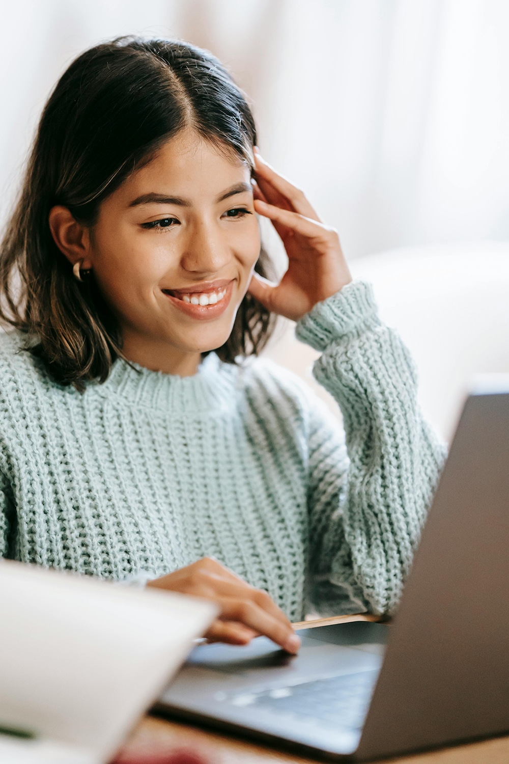 A person with shoulder-length hair smiles while looking at a laptop screen. They are wearing a light blue sweater and sitting indoors.