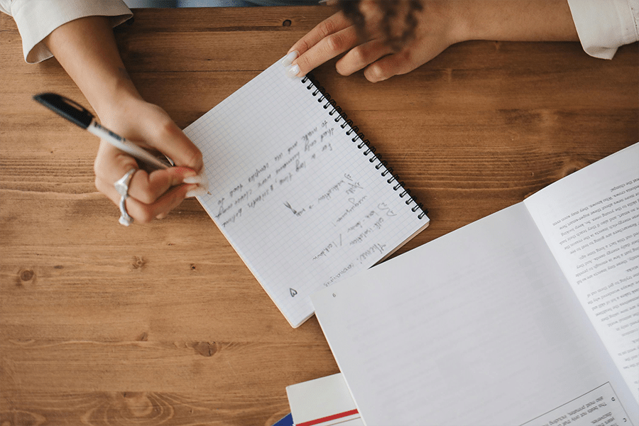 A person writes notes in a notebook on a wooden desk beside an open book and some papers.
