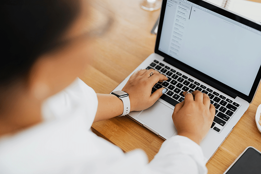 Person wearing a smartwatch typing on a laptop at a wooden desk, with a notebook and pen visible in the background.