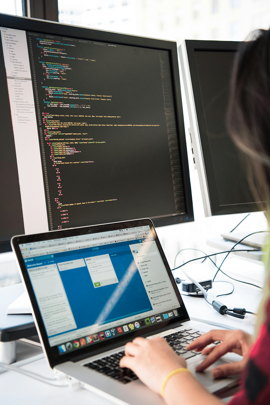 Person typing on a laptop with code displayed on two large monitors in the background.