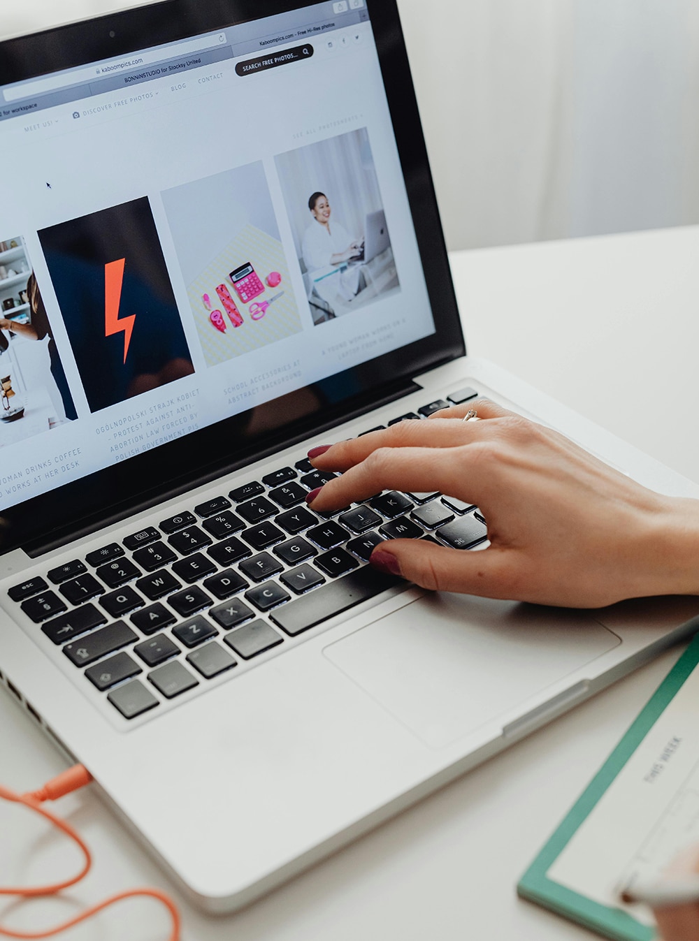 Person using a laptop with an open webpage displaying four images. A notepad and orange cable are next to the laptop.