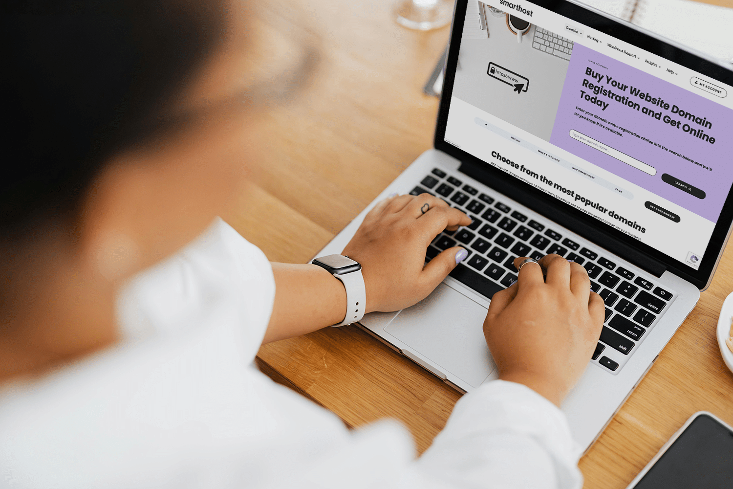 A person types on a laptop, exploring a webpage about buying domain names and how to choose web hosting. A notepad and glasses rest on the desk, ready for jotting down ideas.