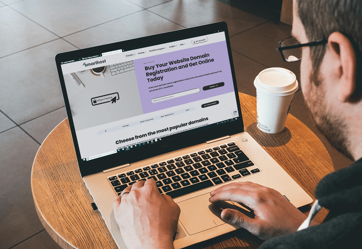 A person sits at a cafe table, their laptop open next to a steaming coffee cup. They are browsing a website for domain registration services, utilizing the domain checker to find the perfect web address for their burgeoning online venture.