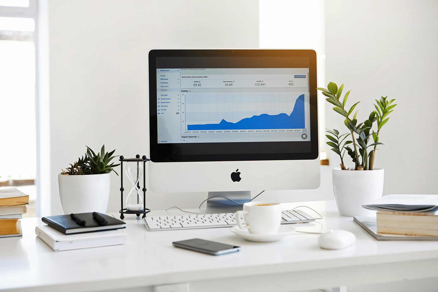 A computer showing a line graph on a desk, flanked by potted plants and notebooks, hints at insights from a comprehensive SEO guide. Nearby, a smartphone, a steaming cup of coffee, and a balance scale complete the scene of productivity and growth.