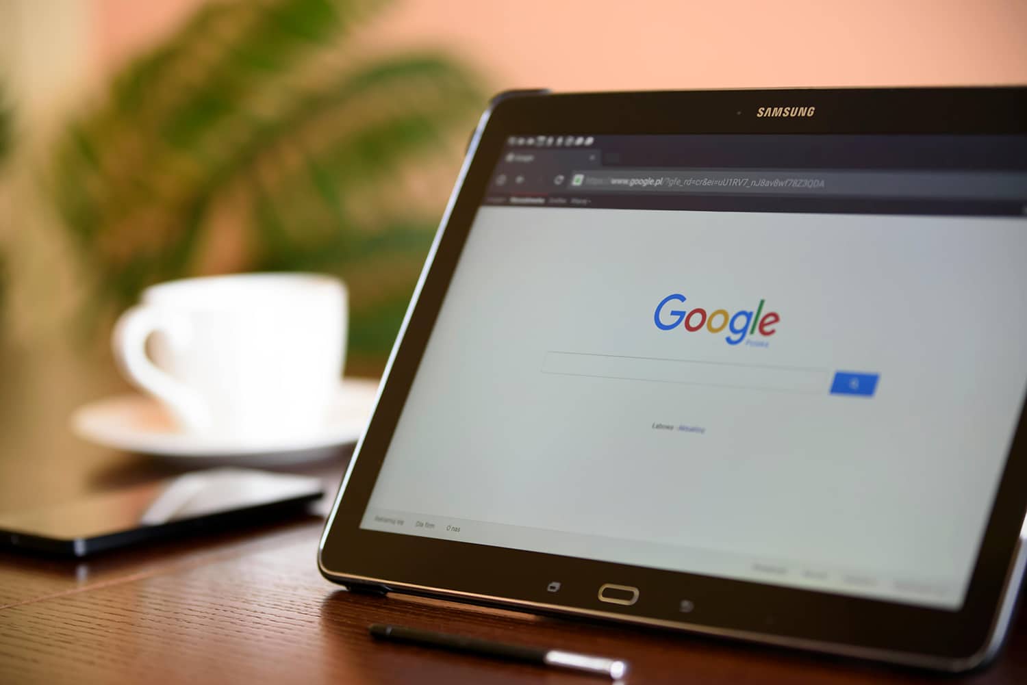 A tablet showcasing the Google search page rests on a table, accompanied by a stylus and smartphone. In the background, a blurred plant and cup enhance the scene. It's as if a comprehensive SEO guide unfolds with every swipe, promising endless exploration and discovery.