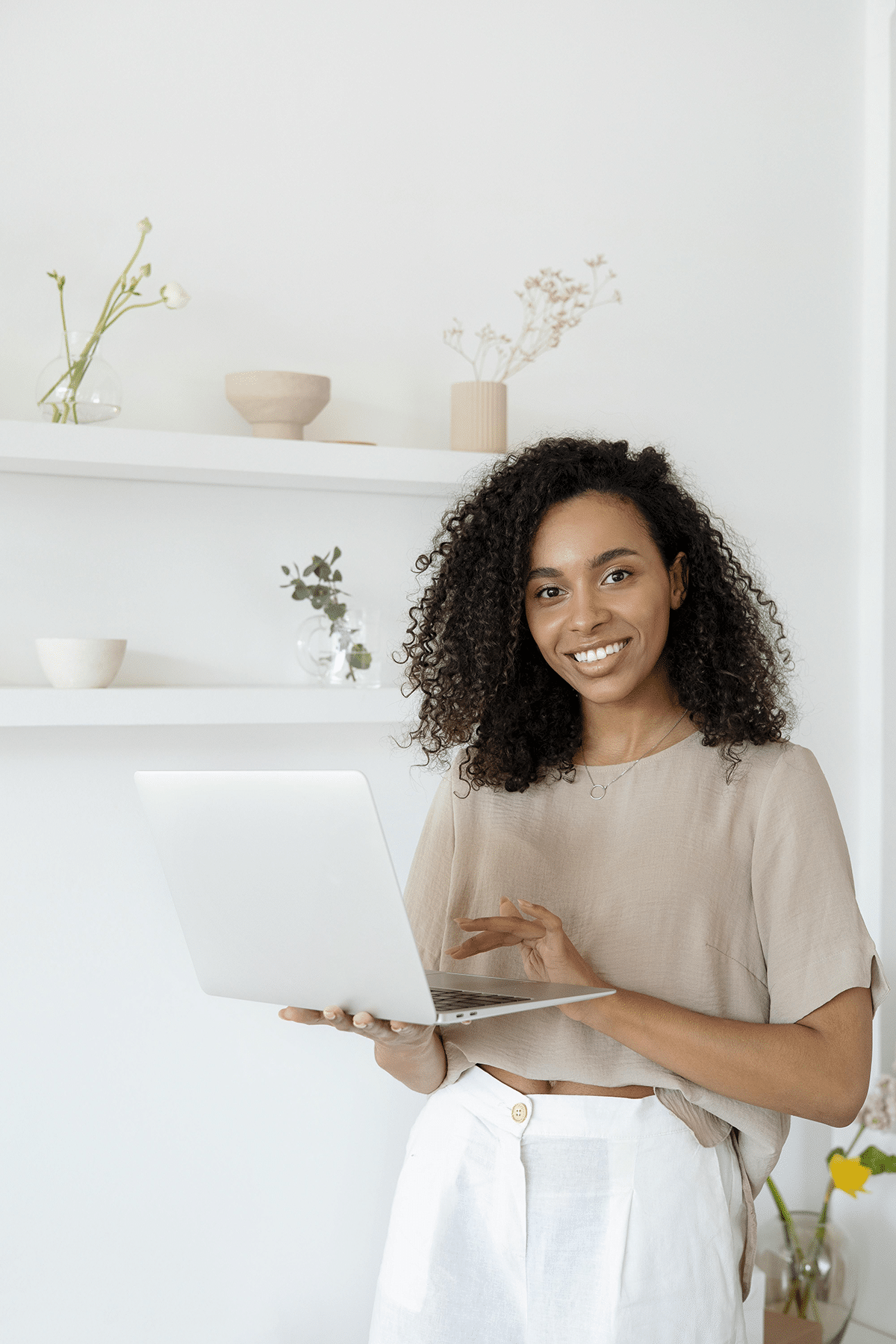 A person with curly hair is smiling while holding a laptop, possibly using a domain checker, in a bright room adorned with shelves and decorative items.