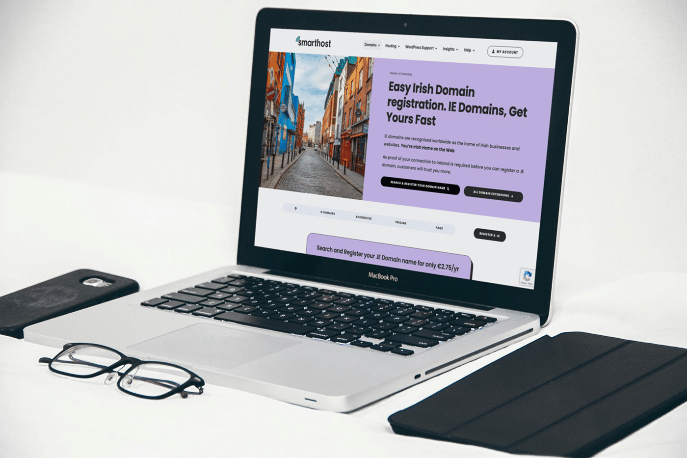A laptop on a desk showcases a website about IE domain registration. Nearby, glasses, a smartphone, and a tablet rest on the pristine white surface.