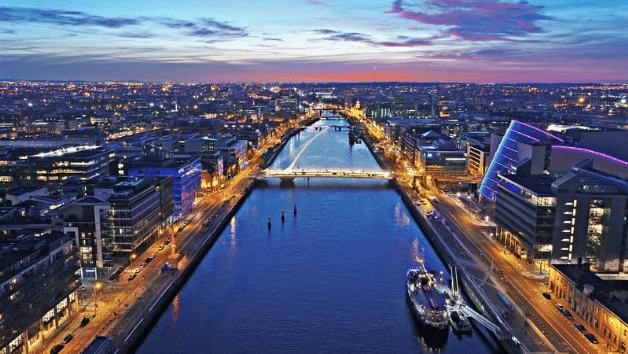 Aerial view of a city river at sunset, where illuminated buildings glow like a guide to learning how to get an IE domain. A bridge gracefully spans the water, with a docked boat in the foreground. The sky displays stunning orange and blue hues.