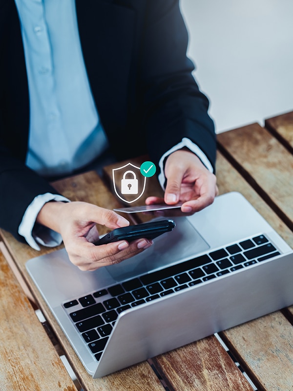 A person in a business suit using a smartphone with an SSL Cert icon above it, while working on a laptop at a wooden table.