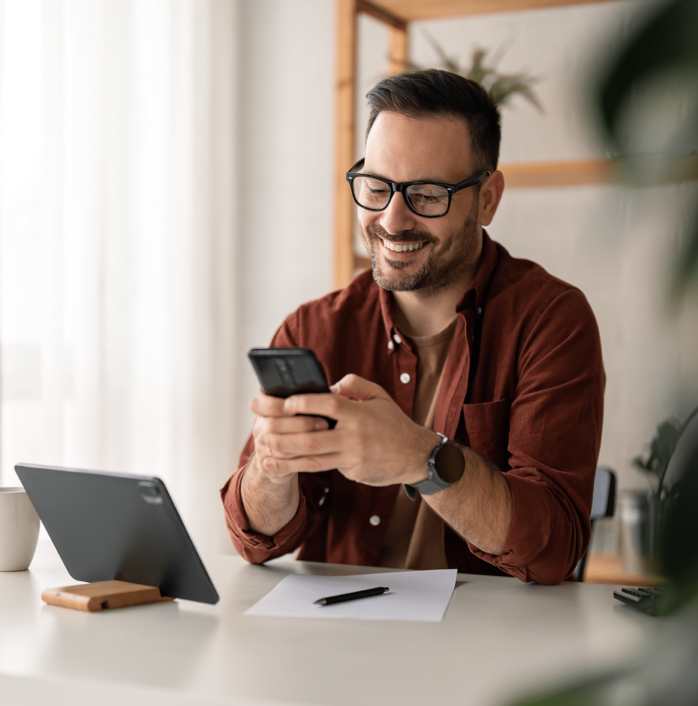 A bearded man in glasses sits at a desk, smiling while using a smartphone. A tablet, paper, pen, and coffee mug are on the desk as he multitasks on technical SEO. Shelves and plants are in the background.
