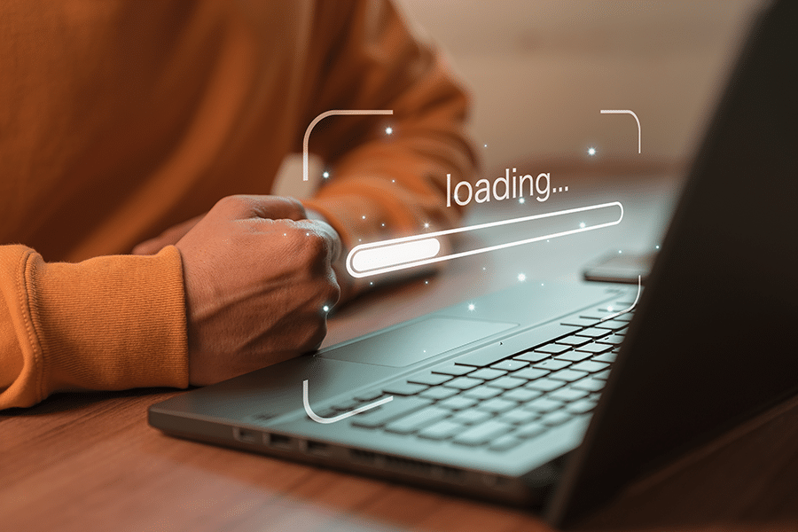 A person in an orange sweater sits at a desk using a laptop. A "loading..." progress bar is superimposed above the laptop keyboard.