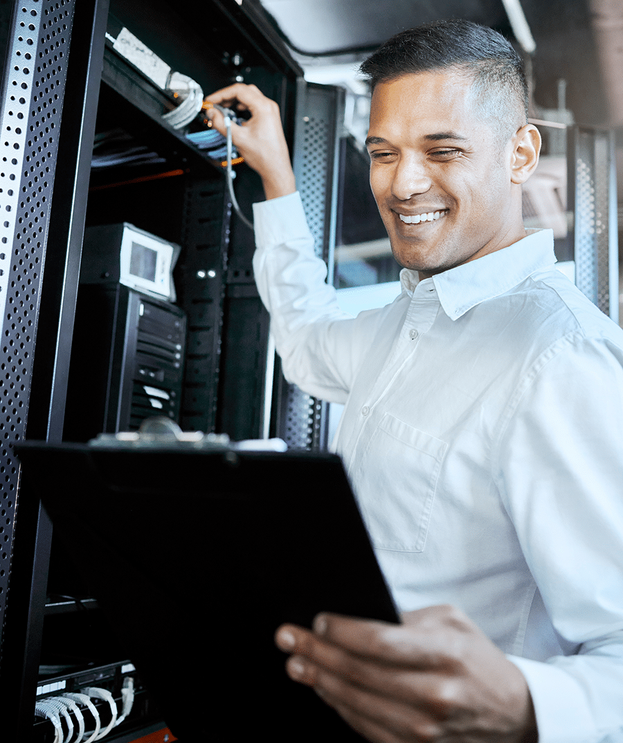 A man in a white shirt, holding a clipboard, checks equipment inside a server rack. He is smiling and appears to be inspecting or maintaining the hardware.