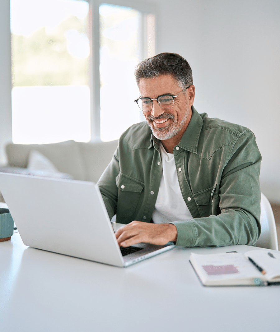 A man wearing glasses and a green shirt works on a laptop at a white desk, near a notebook and a blue cup, in a brightly lit room with large windows.