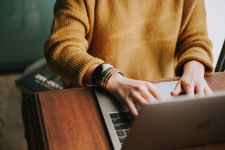 Person in a mustard sweater typing on a laptop at a wooden desk.