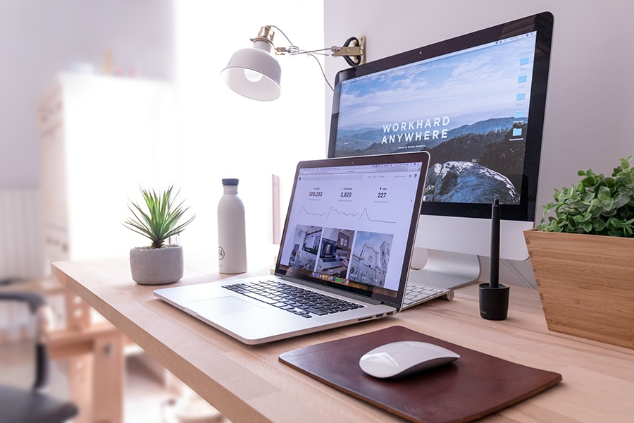 A neatly arranged desk features a laptop, a monitor displaying "Workhard Anywhere," a mouse, a mousepad, a stylus, a water bottle, a potted plant, and a lamp.