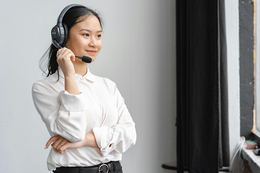 Person wearing a headset with a microphone, standing with folded arms while looking to the side in a white shirt.