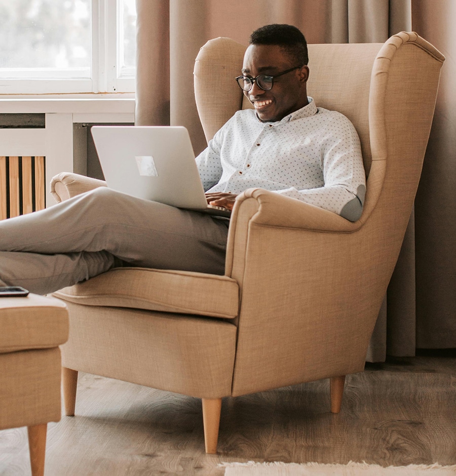 A person sitting in an armchair, smiling and working on a laptop. The setting appears to be a comfortable, well-lit room.