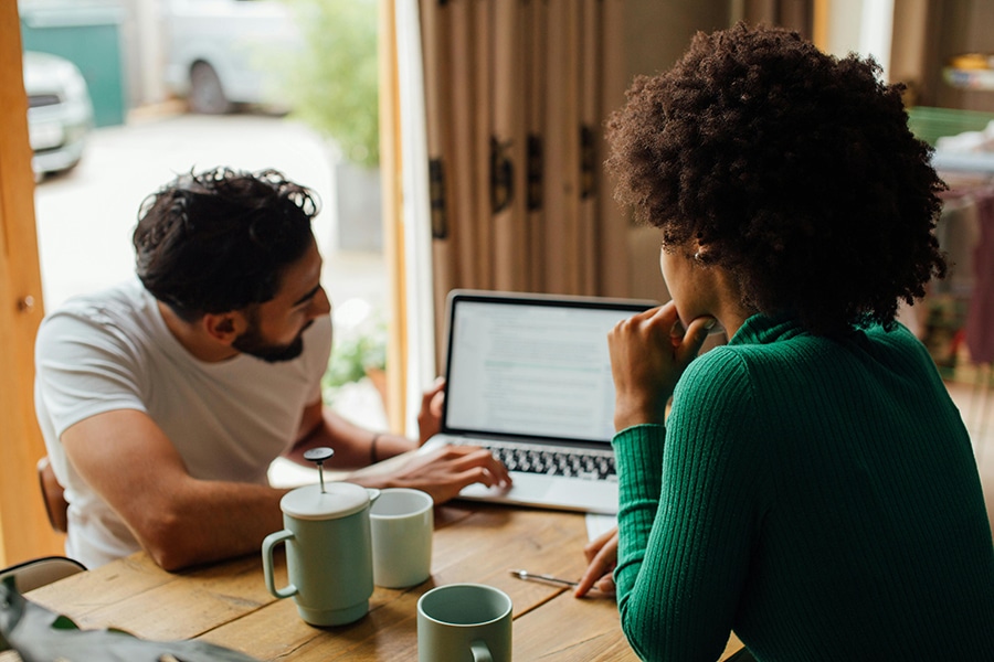 Two people sit at a table with a laptop, focusing intently on the screen. Mugs and a French press are on the table in the cozy, well-lit space as they discuss website speed optimization.