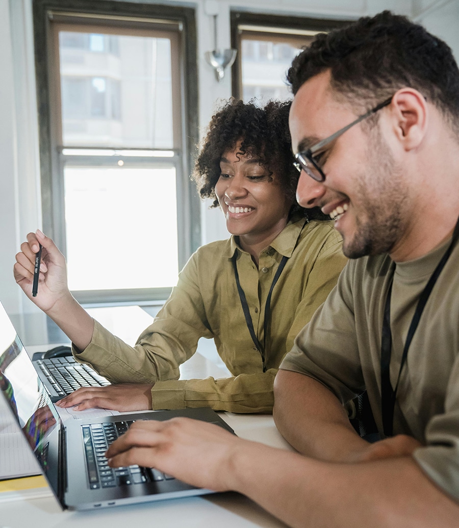 Two people are sitting at a table with laptops, smiling and collaborating in a bright room with large windows as they explore the best strategies for their guide to WordPress Care Plans.