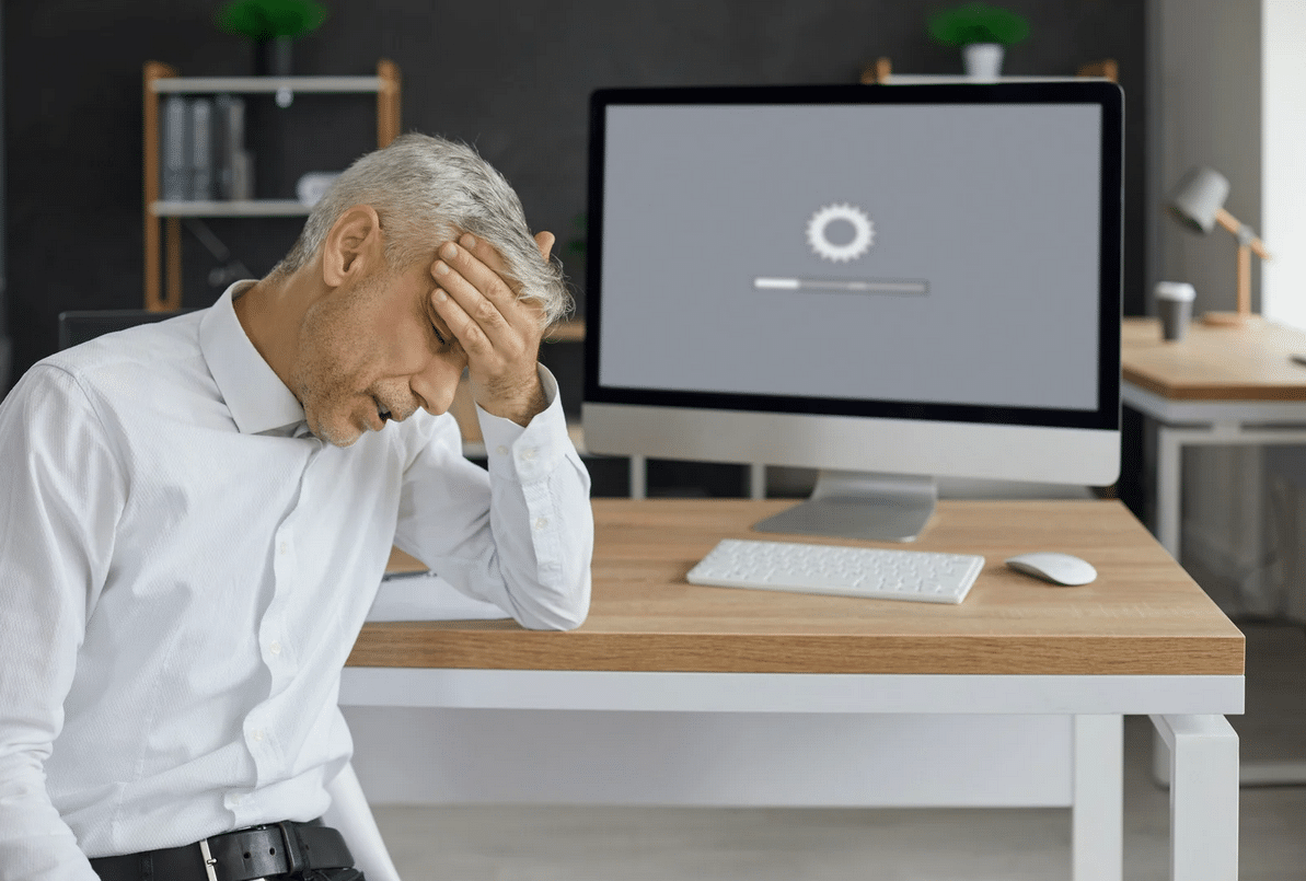A frustrated man in a white shirt holds his head with his hand, seated at a desk with a computer screen displaying a progress bar and gear icon.