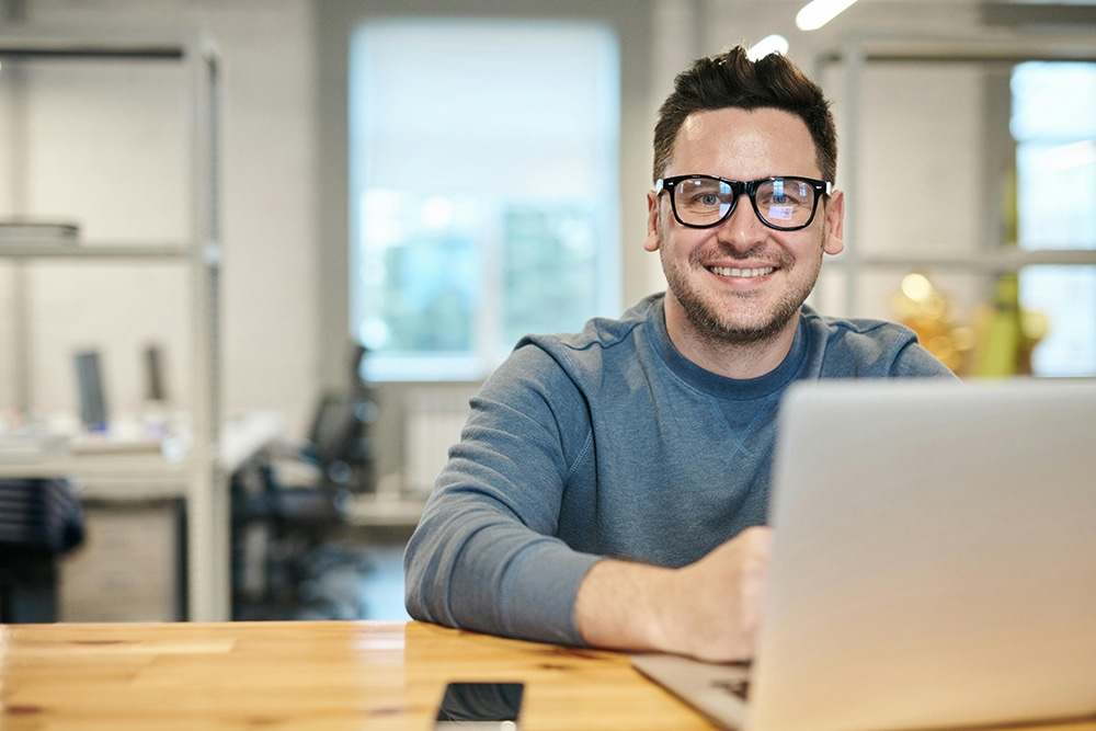 Smiling person wearing glasses and a gray sweater sits at a wooden table using a laptop in a modern office setting.