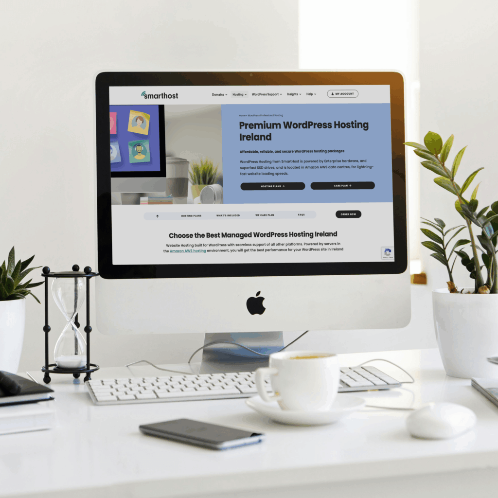 An iMac on a desk displays a website for premium WordPress hosting in Ireland. The workspace also features a keyboard, mouse, phone, hourglass, plant, and a cup—perfect for exploring top Web Hosting Ireland solutions.