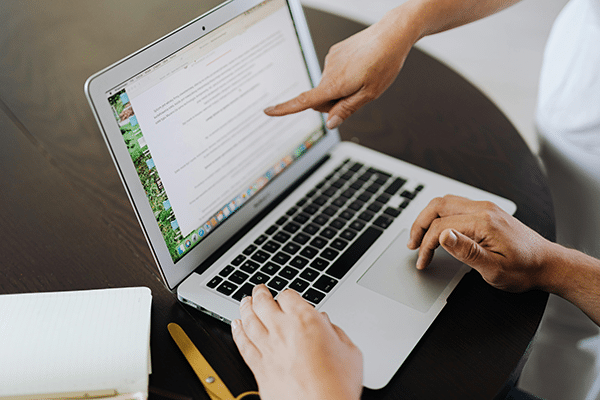 Two people work at a laptop; one types while the other points at text on the screen. A notebook and pen lie on the table beside them.