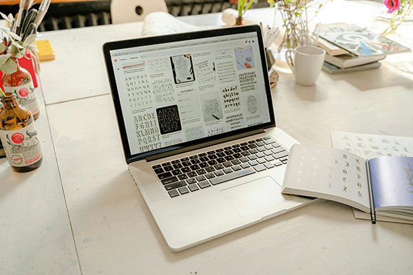 A laptop on a white table displays various fonts and designs. An open notebook, books, bottles, flowers, and a mug are also on the table.