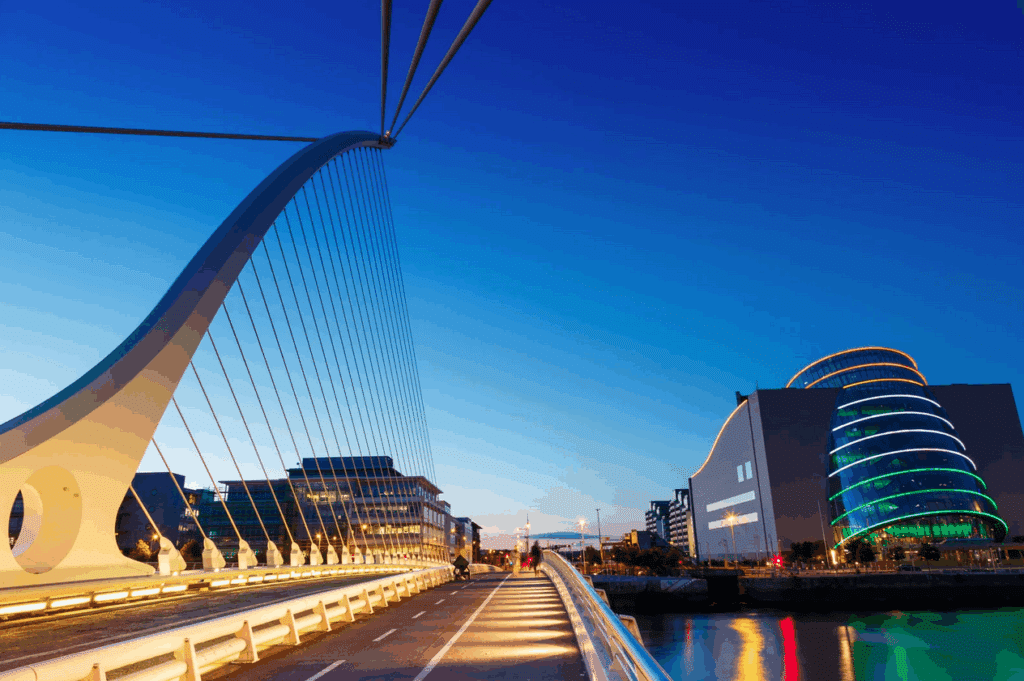 A modern suspension bridge spans a river at dusk, with a curved glass building illuminated with colored lights on the right.
