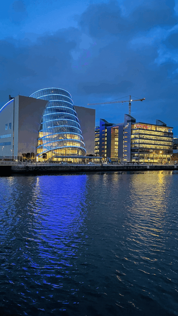 Modern glass buildings with bright lights reflecting on the water at dusk, under a cloudy blue sky. A crane is visible in the background.