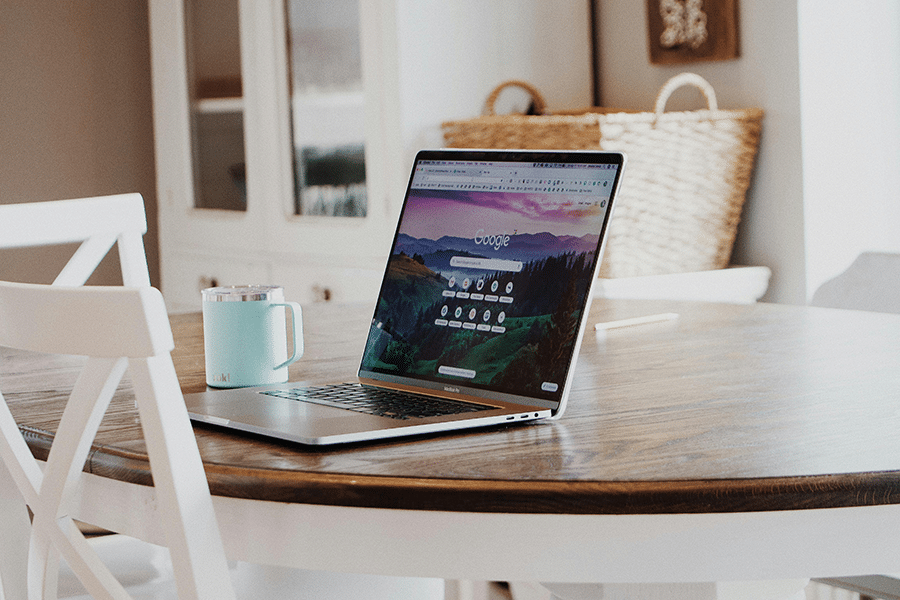 A laptop displaying a search engine homepage sits on a round wooden table next to a light blue mug in a bright, cozy room.