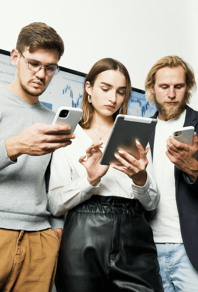 Three people standing close together, looking at electronic devices; two using smartphones and one using a tablet, with a digital screen in the background.