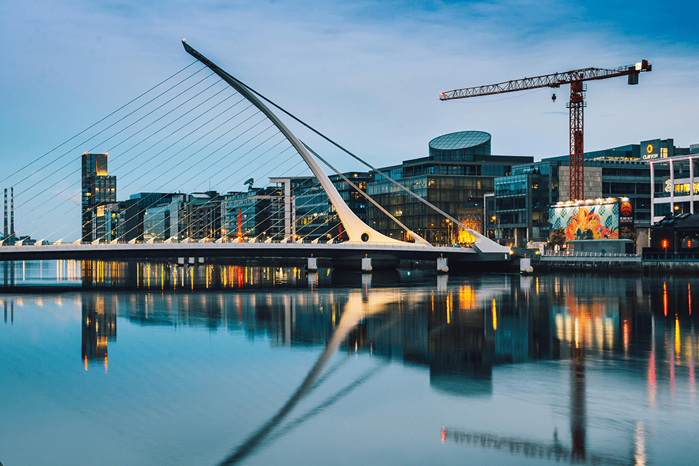 A modern cable-stayed bridge spans a calm river, with city buildings, a crane, and colorful mural visible in the background at dusk.