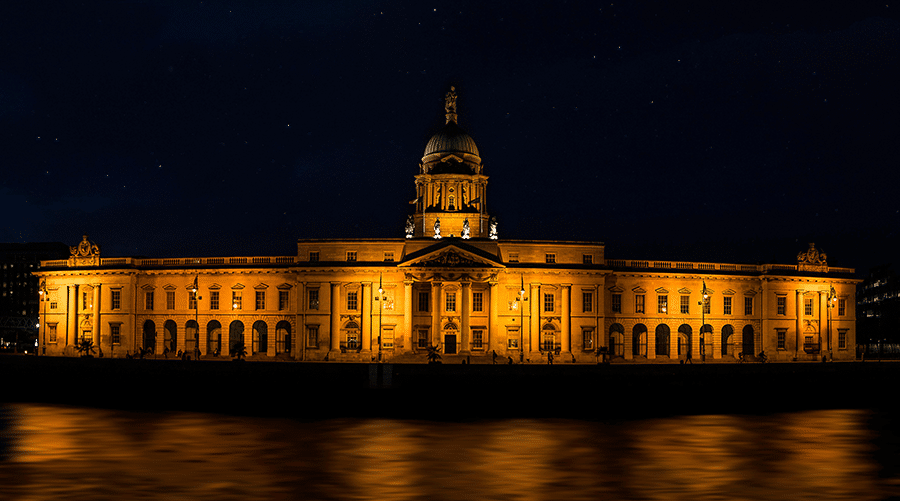 A large neoclassical building with a dome, illuminated by yellow lights at night, stands beside a calm body of water.