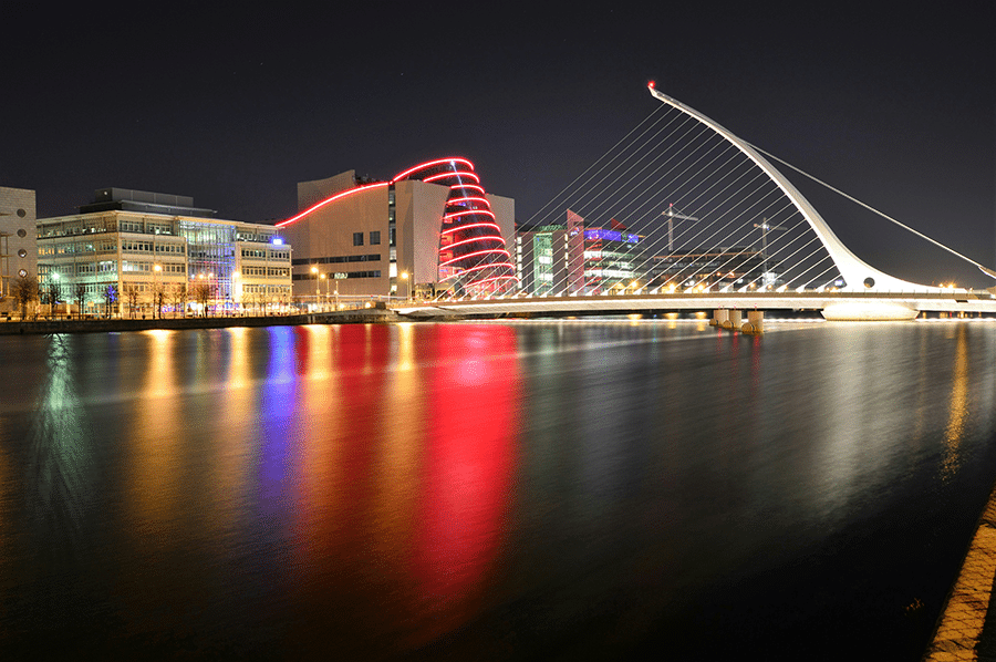 A night view of the Samuel Beckett Bridge and illuminated buildings reflecting on the River Liffey in Dublin, Ireland.