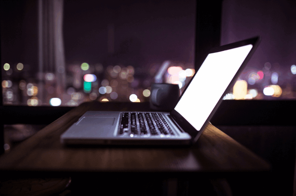 A laptop with a bright screen sits on a wooden table next to a mug, with city lights visible through a window in the background at night.