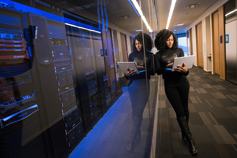 A woman stands in a hallway next to server racks, holding a laptop and looking at the screen, with her reflection visible in the glass wall beside her.