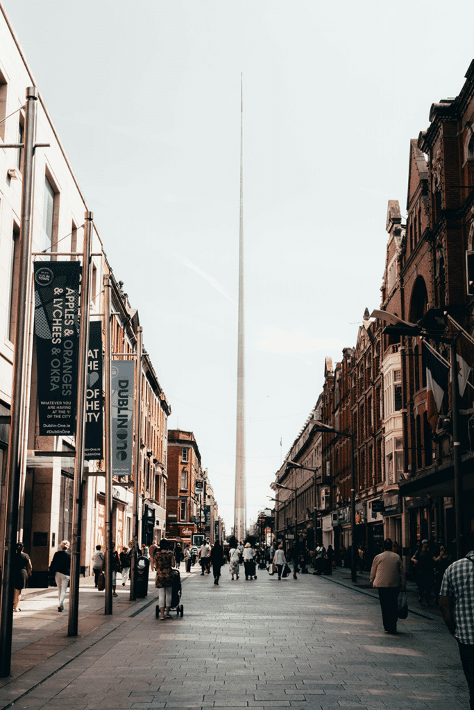 Pedestrians walk along a city street lined with shops and historic buildings, with a tall, pointed monument (the Spire of Dublin) rising in the center background.