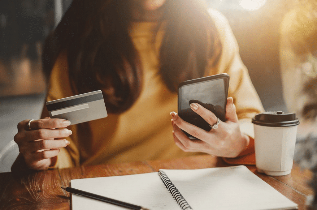 A person holding a credit card and a smartphone sits at a wooden table with a notebook and a takeaway coffee cup.