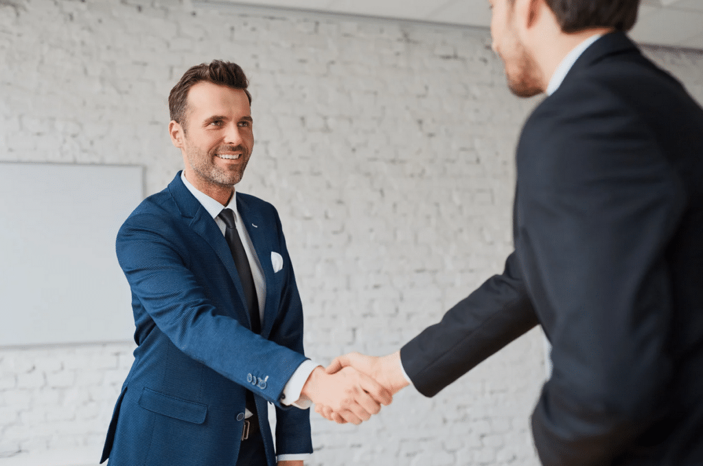 Two men in business suits shake hands in a modern office with a white brick wall in the background.